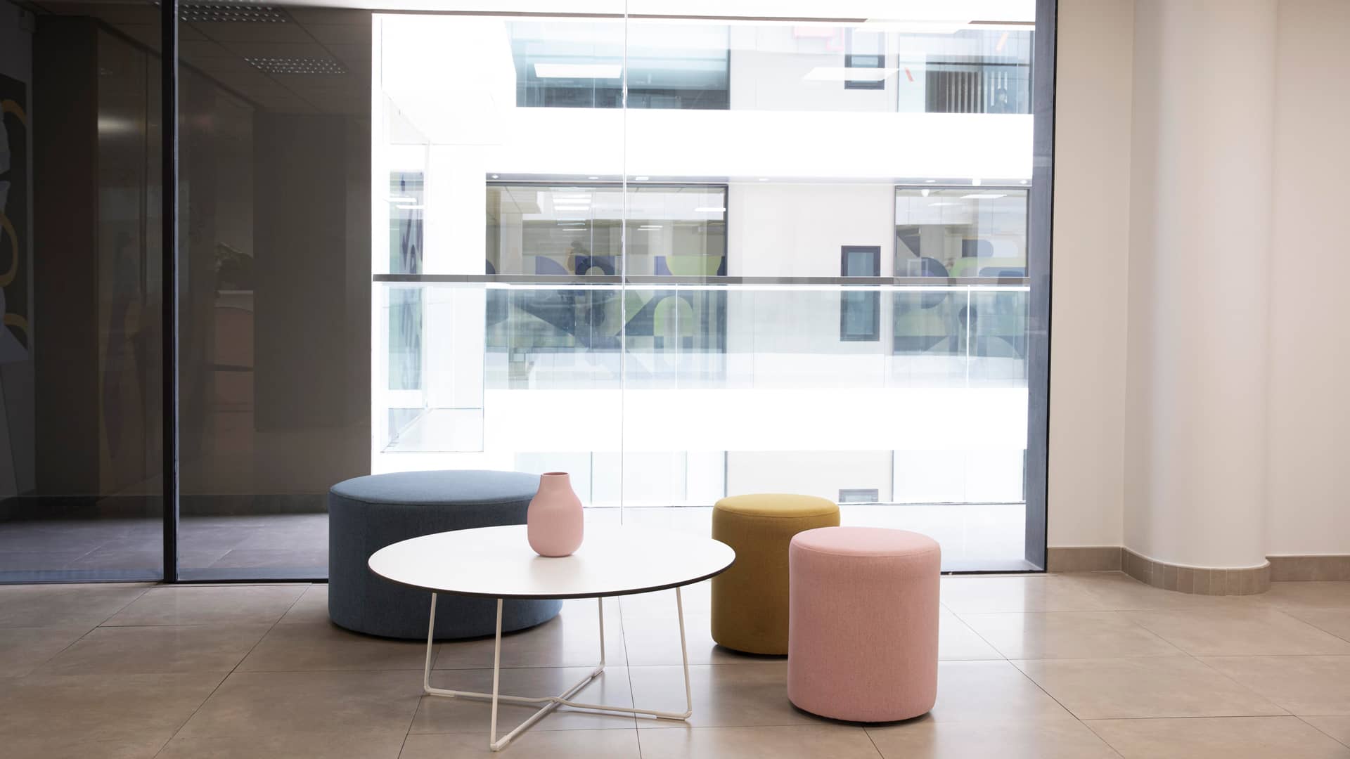 Ein moderner Warte- oder Lounge-Bereich mit einem runden weißen Tisch, auf dem eine kleine Vase steht. Um den Tisch herum stehen drei gepolsterte Hocker in Blau, Gelb und Rosa. Im Hintergrund sind große Fenster mit Blick auf ein modernes Gebäude mit Glasbalkonen.