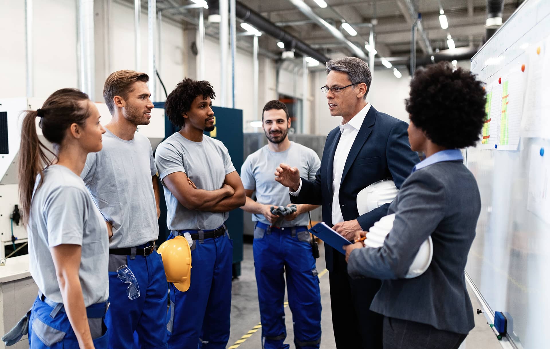 Ein Team von Handwerkerinnen und Handwerkern in blauer Kleidung hört aufmerksam einem Chef in Anzug zu. Der Chef hält einen weißen Helm und spricht mit klaren Gesten. Alle wirken interessiert und konzentriert. Im Hintergrund sieht man Maschinen und eine helle Werkstatt.