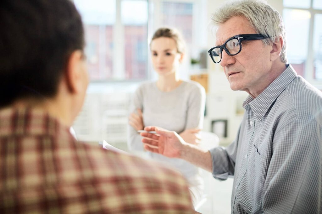 Die Abbildung zeigt drei Personen in einem Gespräch in einem hellen Büro. Ein älterer Mann mit grauen Haaren, Brille und grauem Hemd spricht gestikulierend mit zwei anderen Personen. Eine Frau mit zusammengebundenen Haaren trägt ein helles Oberteil und hört aufmerksam zu, während eine dritte Person, die teilweise von hinten zu sehen ist, ebenfalls an dem Gespräch teilnimmt. Das Umfeld ist modern und gut beleuchtet.